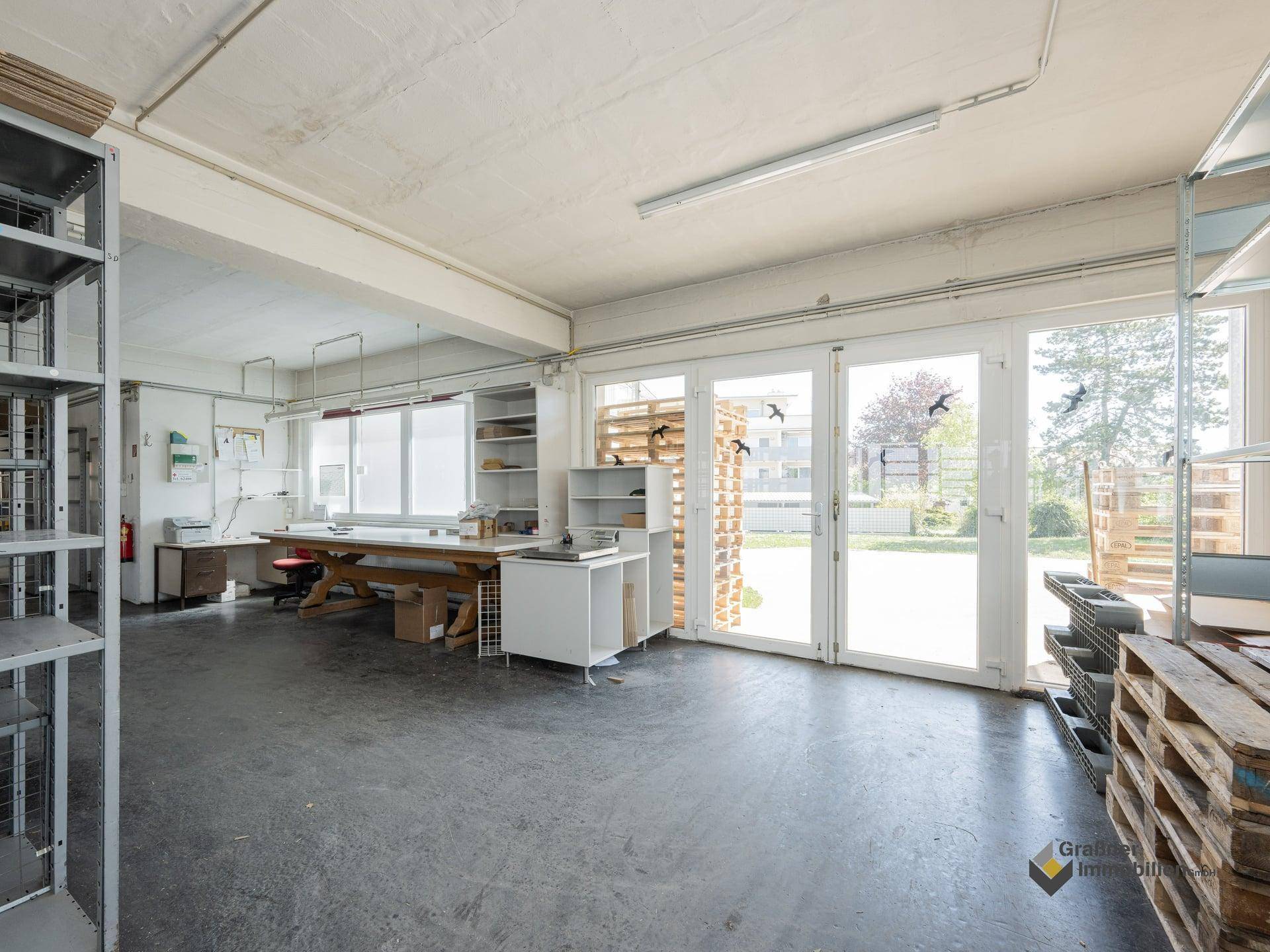 Geräumiger Werkstattraum mit Betonboden, Regalen und großen Glastüren zum Außenbereich.
