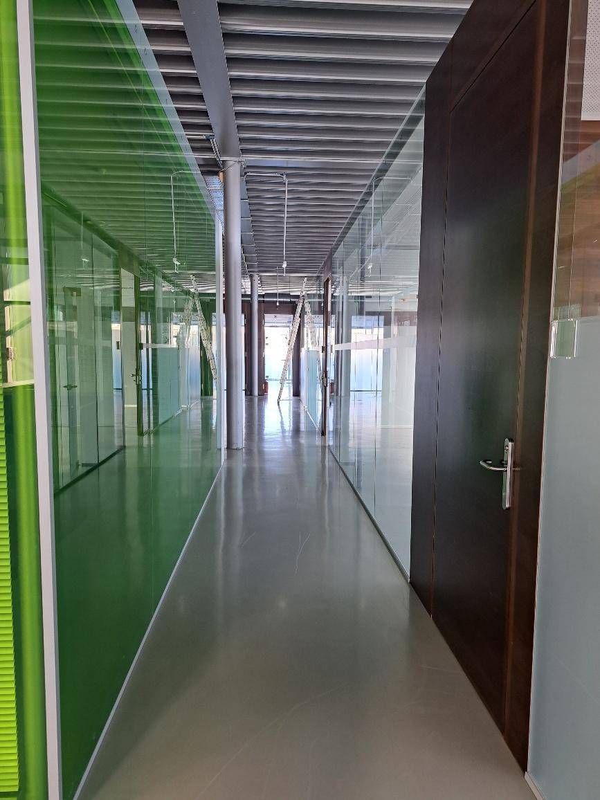 Moderner Flur mit Glaswänden, die Büros abtrennen, und einem glänzenden Betonboden, der Helligkeit reflektiert.