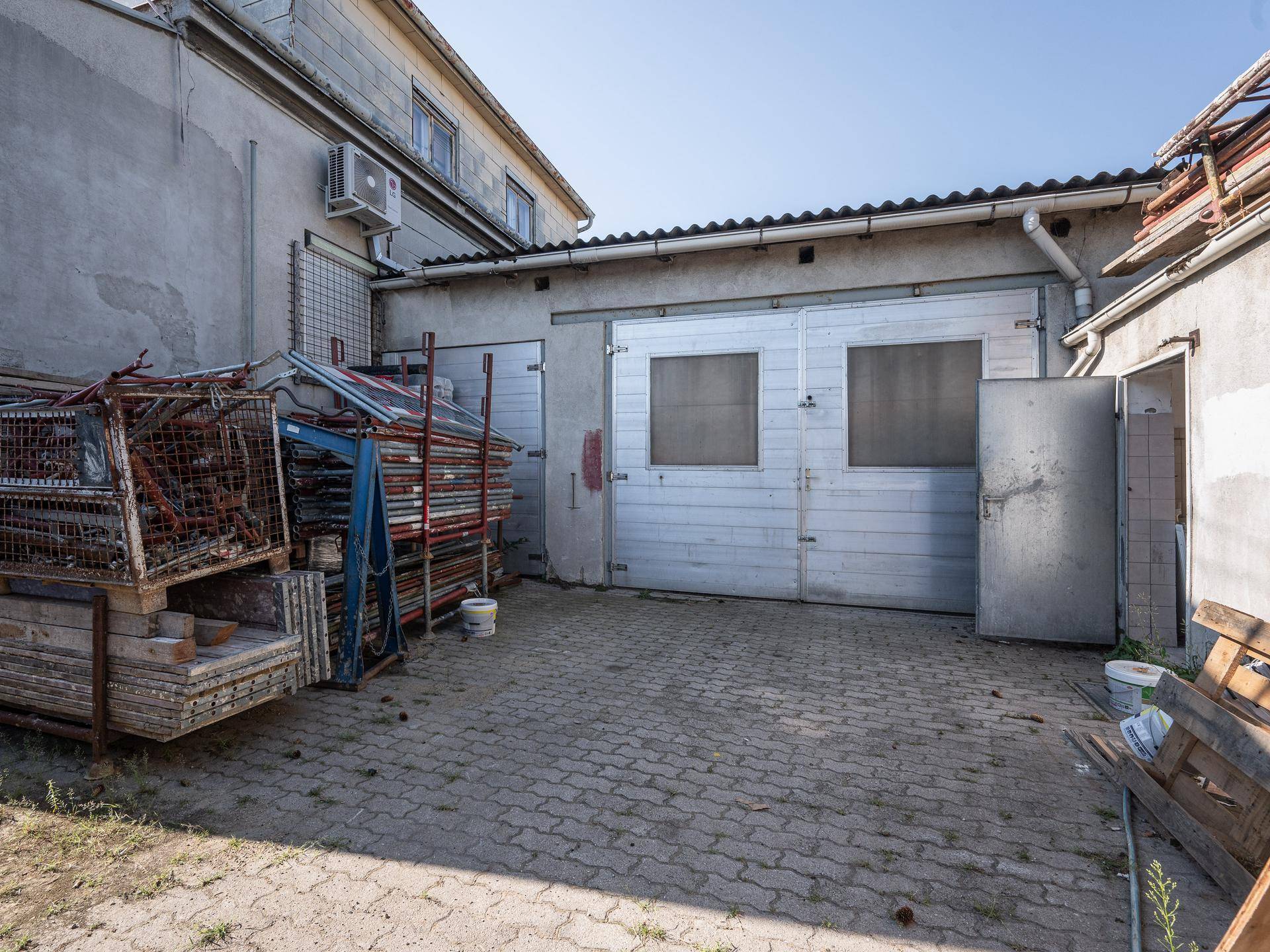 Außenansicht eines Gebäudes mit zwei Garagentoren und einem offenen Seiteneingang, umgeben von Baumaterialien.