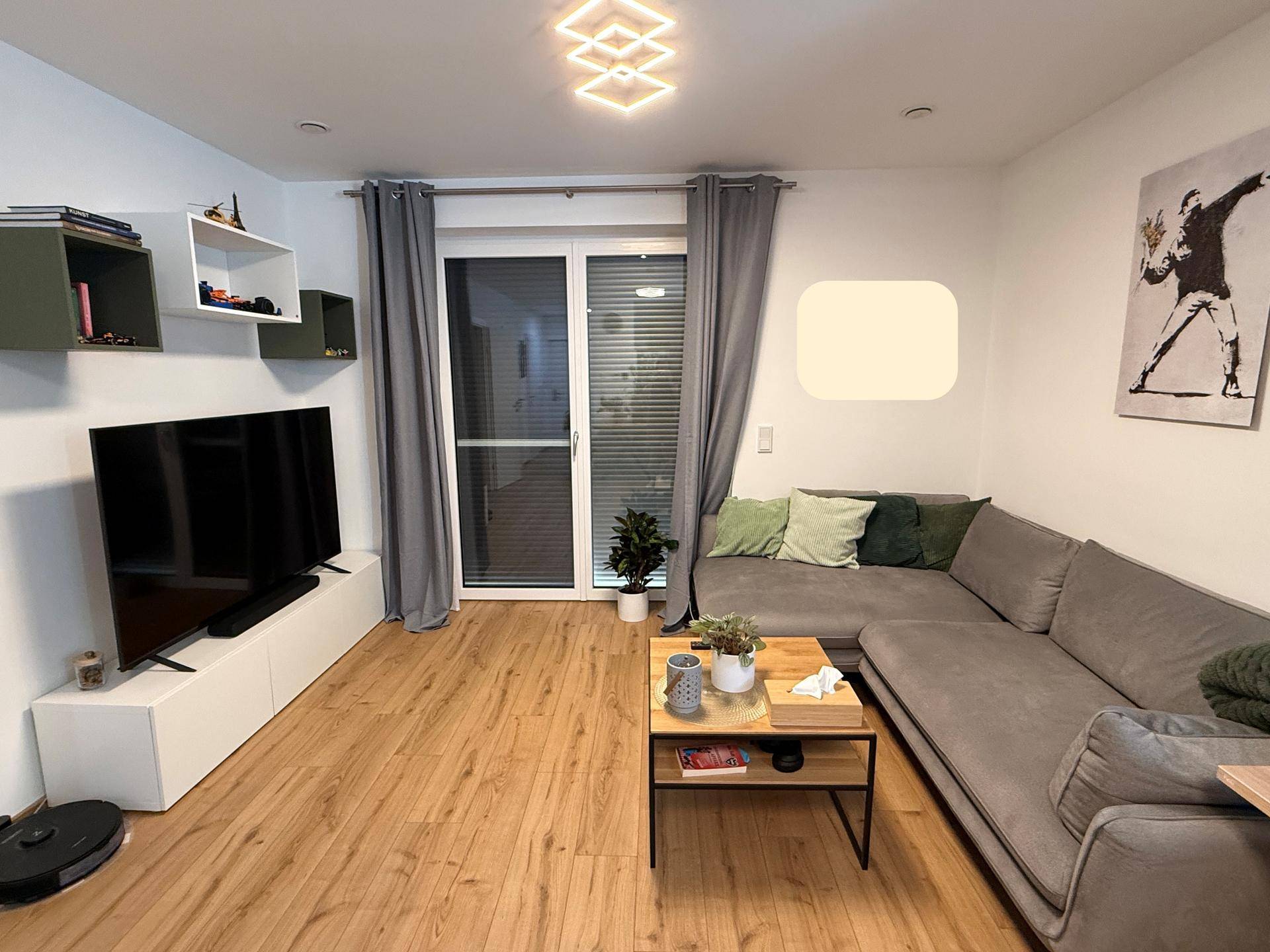 Gemütliches Wohnzimmer mit grauem Sofa, Fernseher und Zugang zu einem Außenbereich.