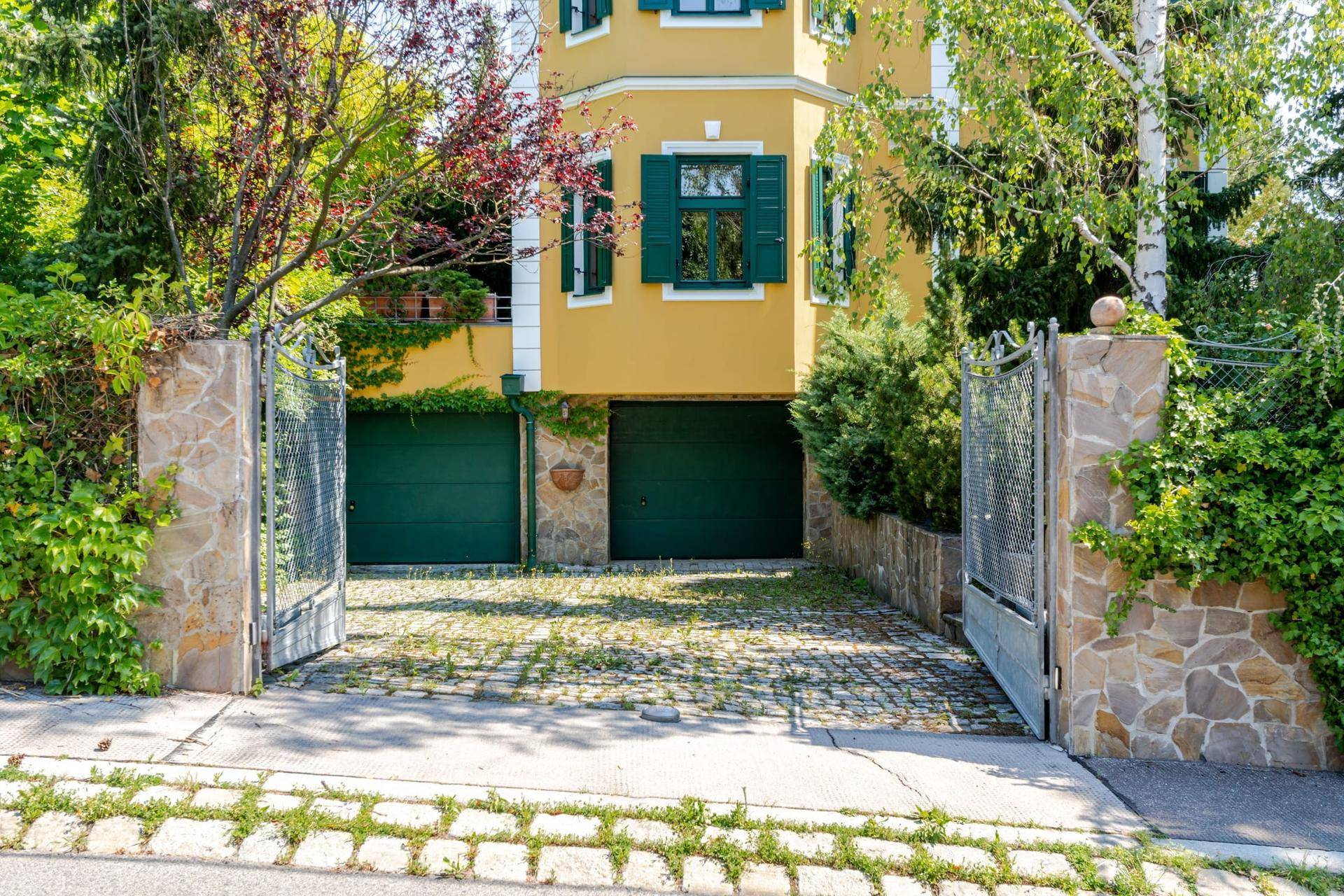 Eingangsbereich der Villa mit zwei Garagentoren und gepflasterter Auffahrt, umgeben von Grün.