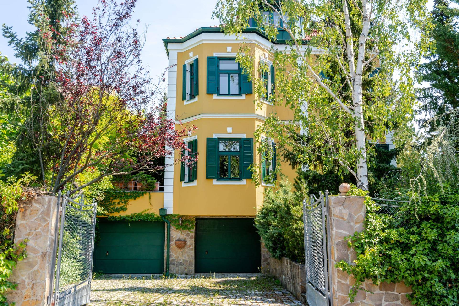 Ansicht der Villa mit gelber Fassade, grünen Fensterläden und zwei Garagenzugängen.