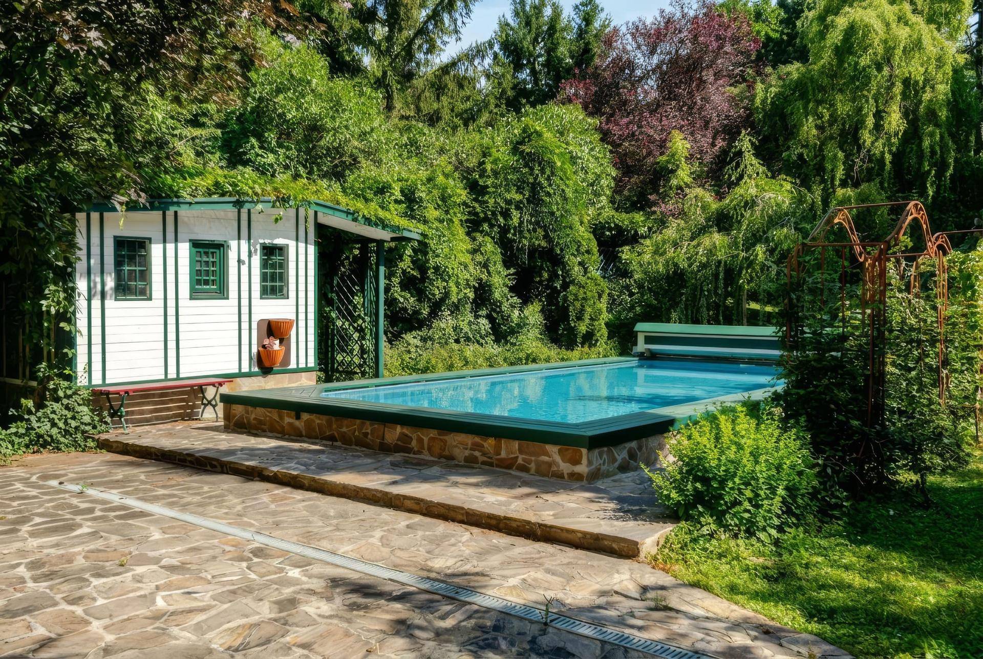 Außenpoolbereich mit kleinem Poolhaus und gepflastertem Weg, umgeben von üppigem Grün.