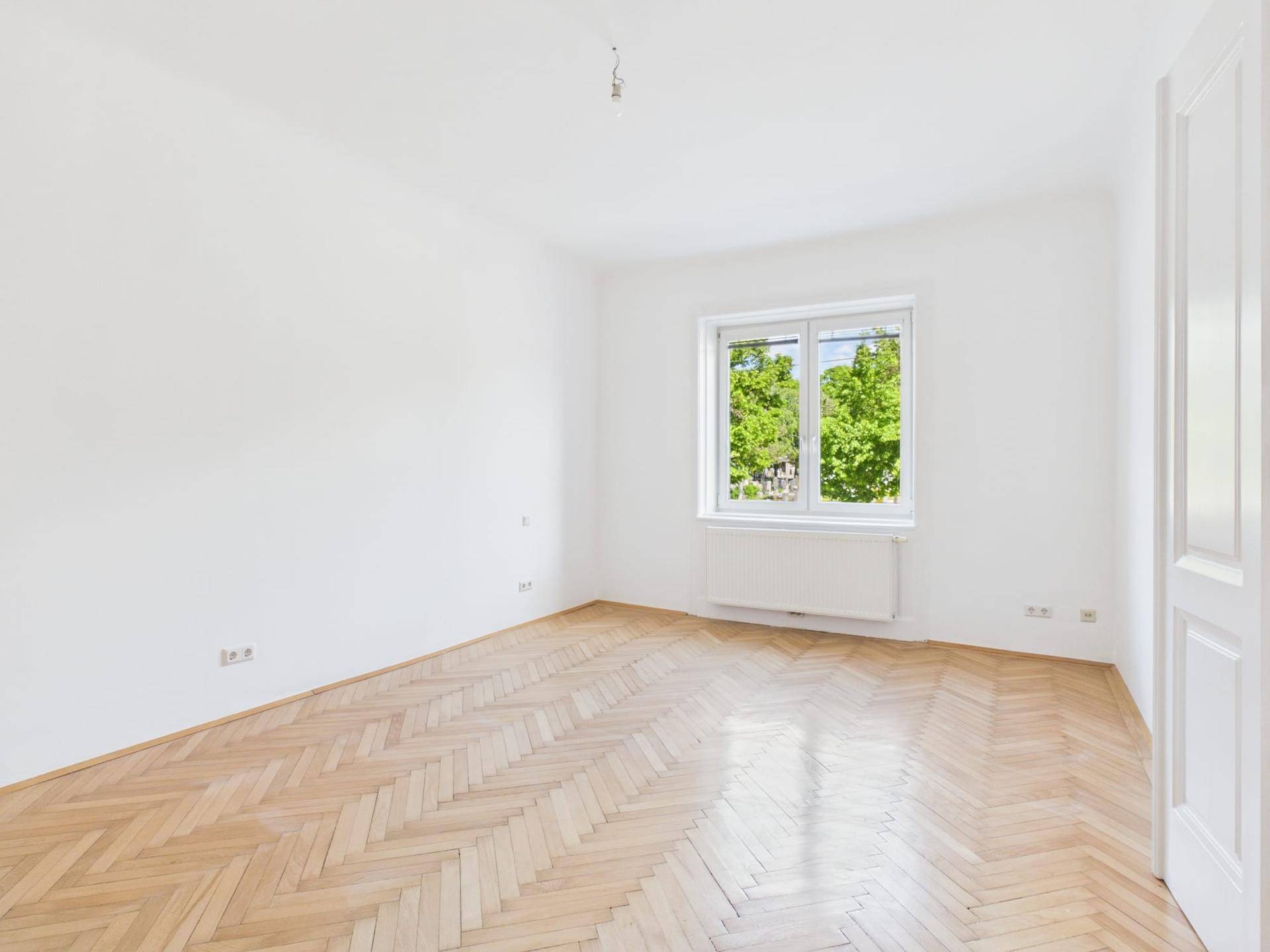Heller, leerer Raum mit Fischgrätparkettboden und Blick ins Grüne durch das Fenster.