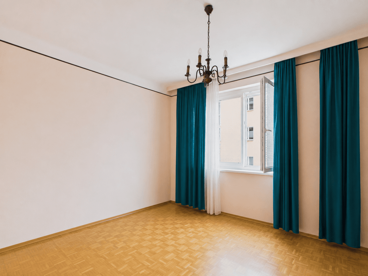 Geräumiges, leeres Zimmer mit Parkettboden, Kronleuchter und geöffnetem Fenster mit blauen Vorhängen.