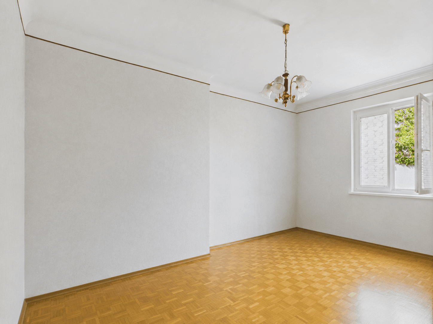 Helles, leeres Zimmer mit Parkettboden und einem Fenster, das viel Tageslicht hereinlässt.
