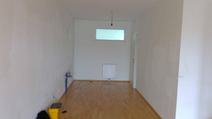 Leerer Raum mit hellem Parkettboden, weißen Wänden und einem kleinen Fenster, vorbereitet für Installationen.