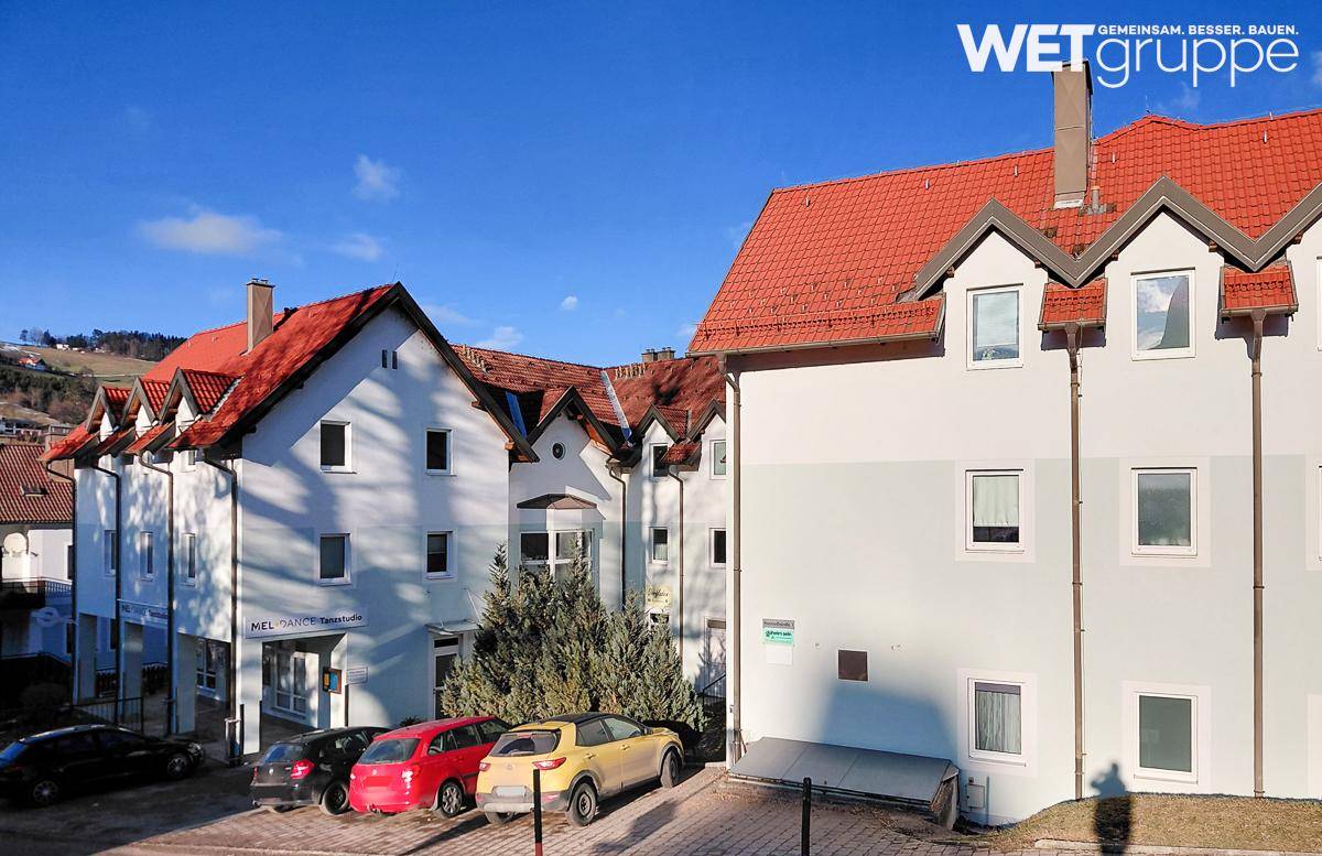Wohngebäude mit roten Ziegeldächern und Parkplätzen, umgeben von grüner Landschaft unter blauem Himmel.