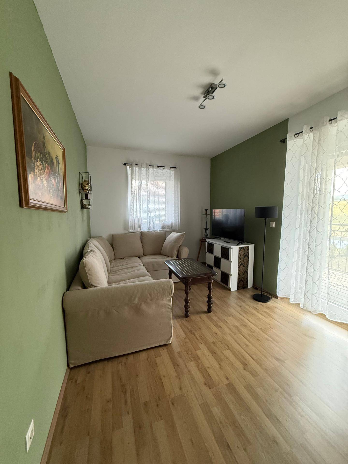 Helles Wohnzimmer mit beiger Eckcouch, grün gestrichener Wand und Laminatboden.