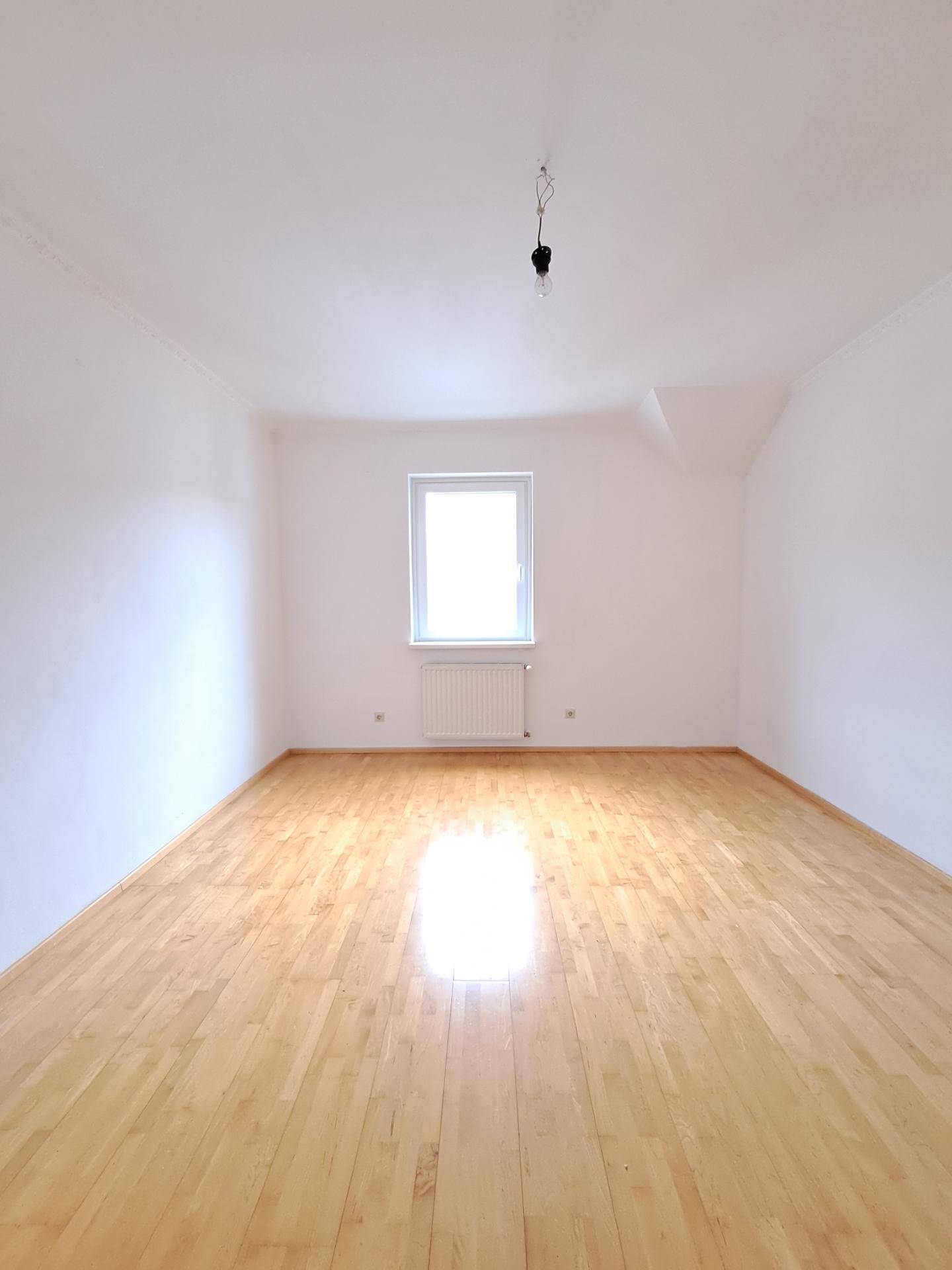 Leerer, heller Raum mit Laminatboden und einem Fenster, das viel Tageslicht in den Raum lässt.