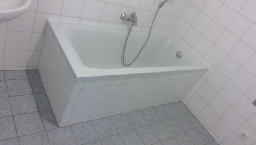 Badezimmer mit weiß gefliester Badewanne, weißen Wandfliesen und grauen Bodenfliesen.