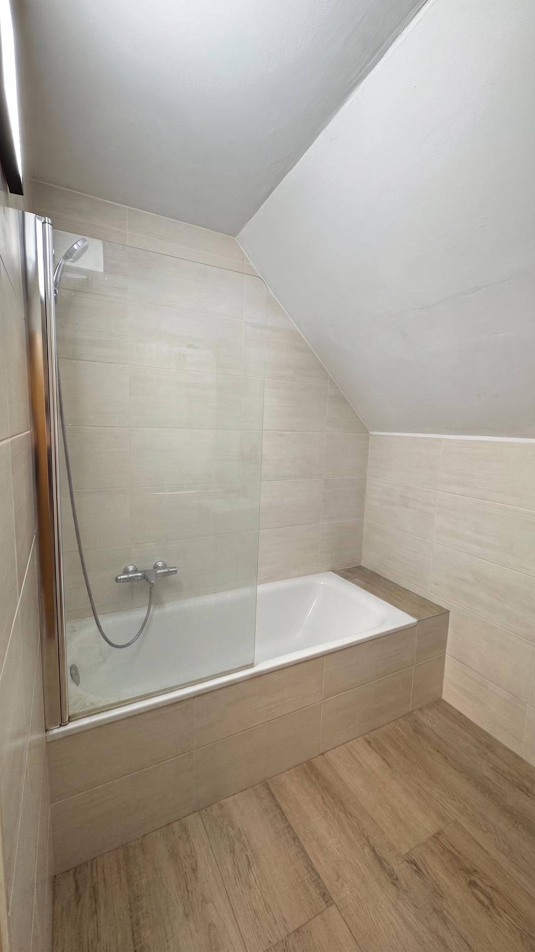 Badezimmer im Dachgeschoss mit Badewanne, Duschabtrennung und beigefarbenen Fliesen.