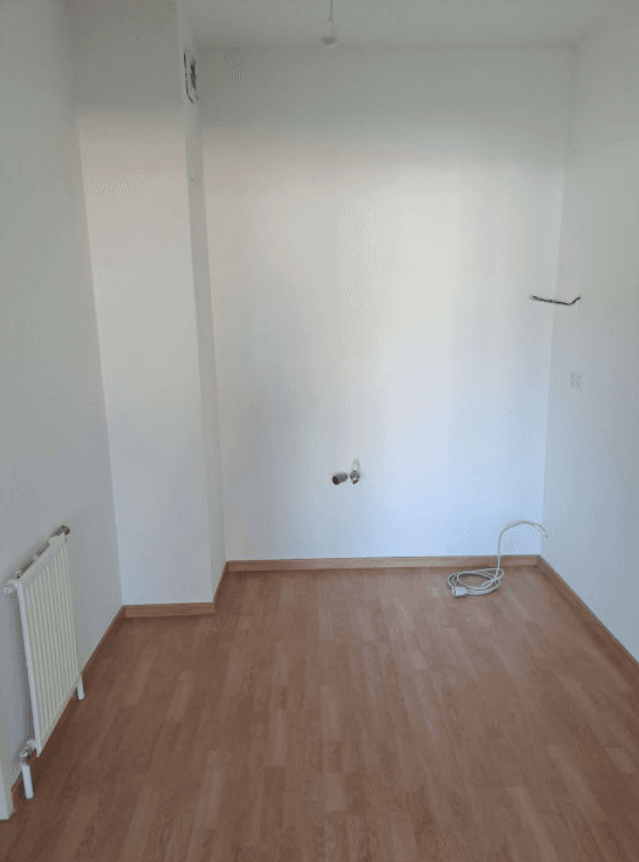 Leerer Küchenraum mit Laminatboden, Heizkörper und Anschlüssen für eine zukünftige Einbauküche.