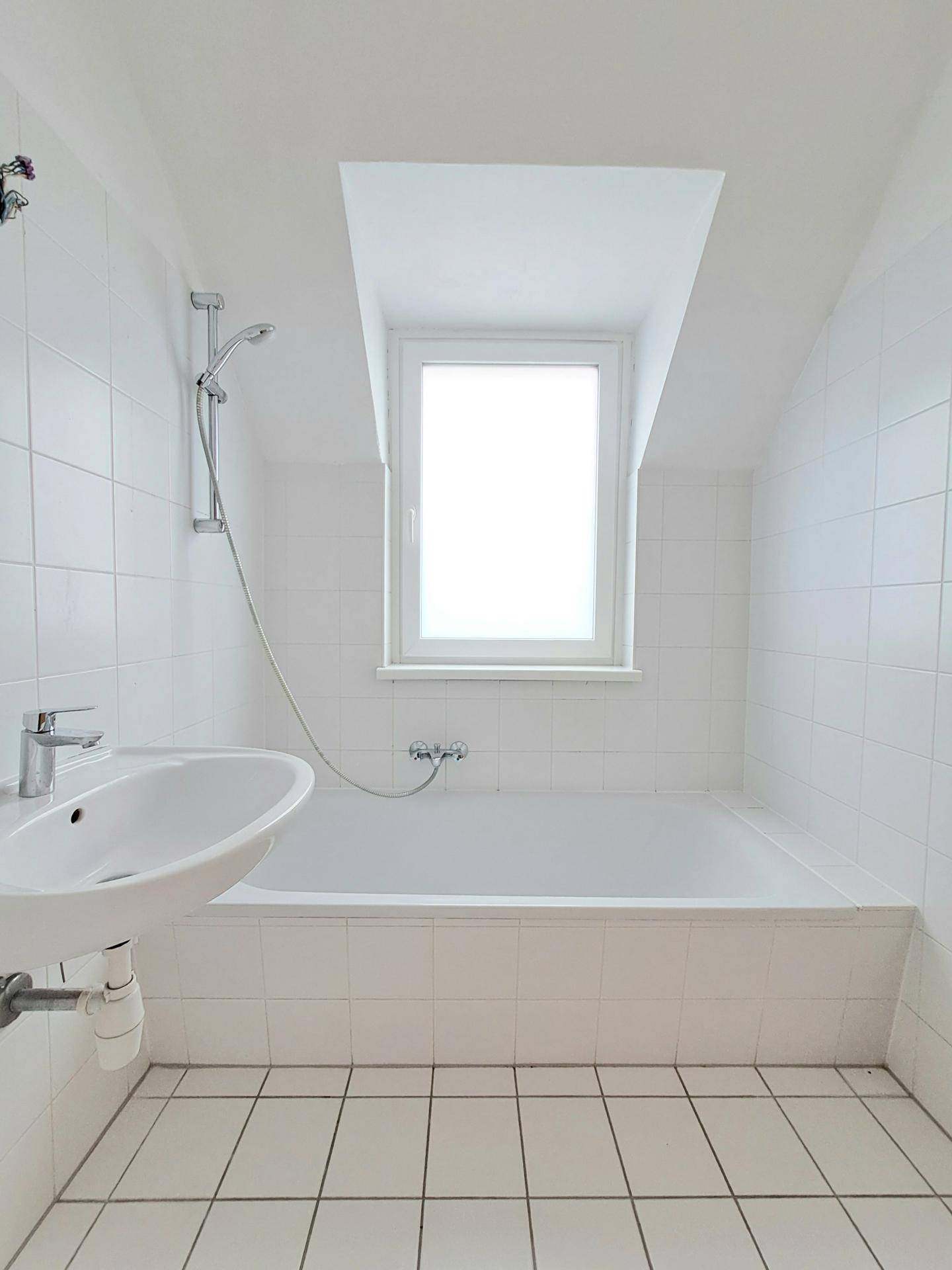 Helles Badezimmer mit weißen Fliesen, Badewanne und Fenster unter einer Dachschräge, in gutem Zustand.