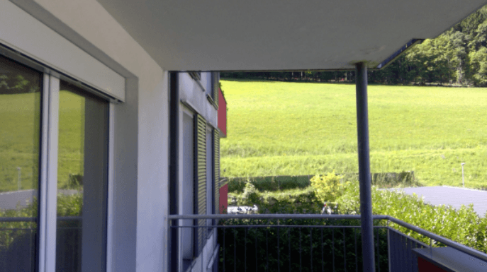 Balkon mit Blick auf eine weite grüne Wiese und bewaldete Hügel im Hintergrund.