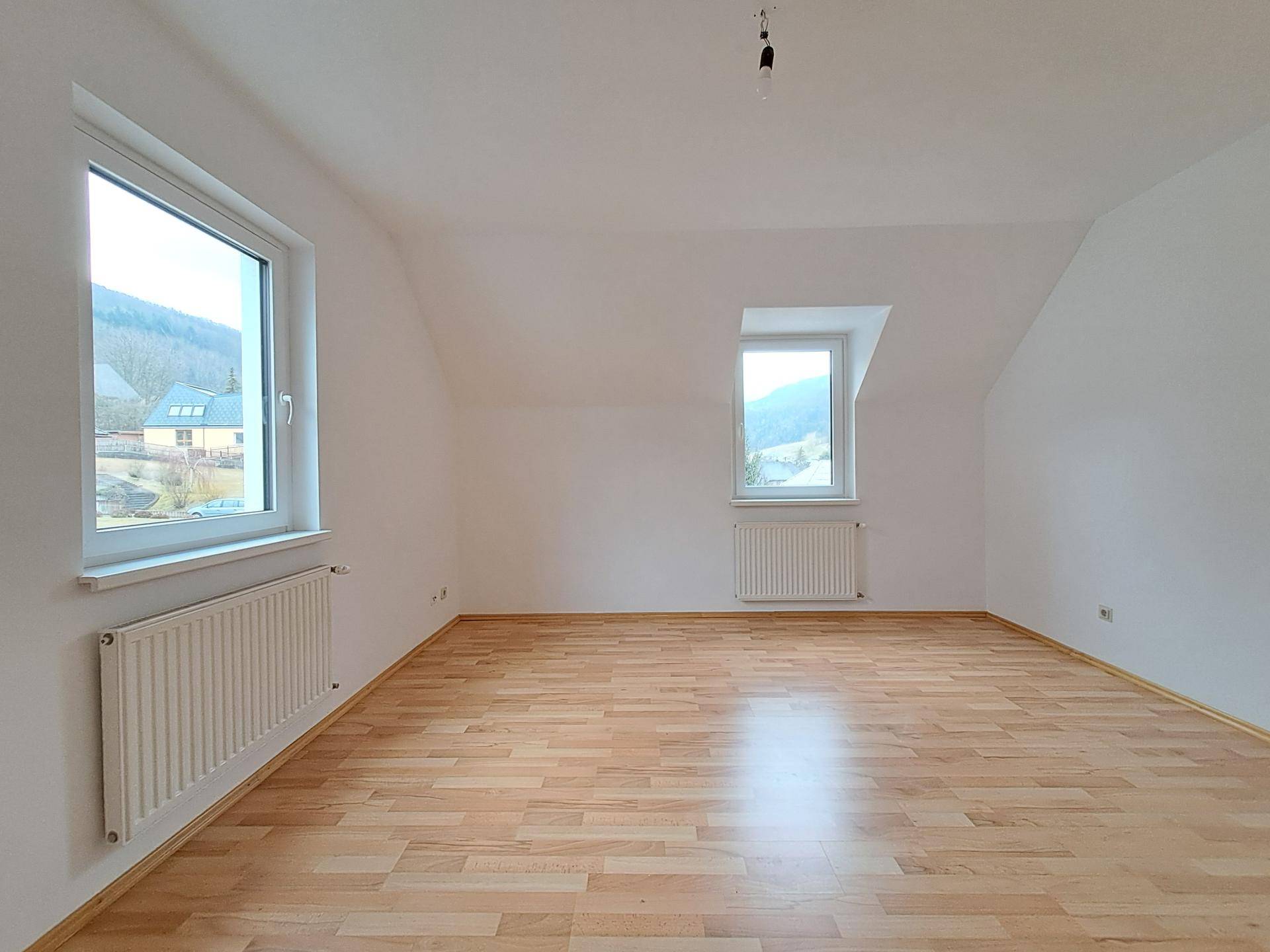 Heller, leerer Raum mit Laminatboden, Dachschrägen und zwei Fenstern, die einen Blick ins Grüne bieten.