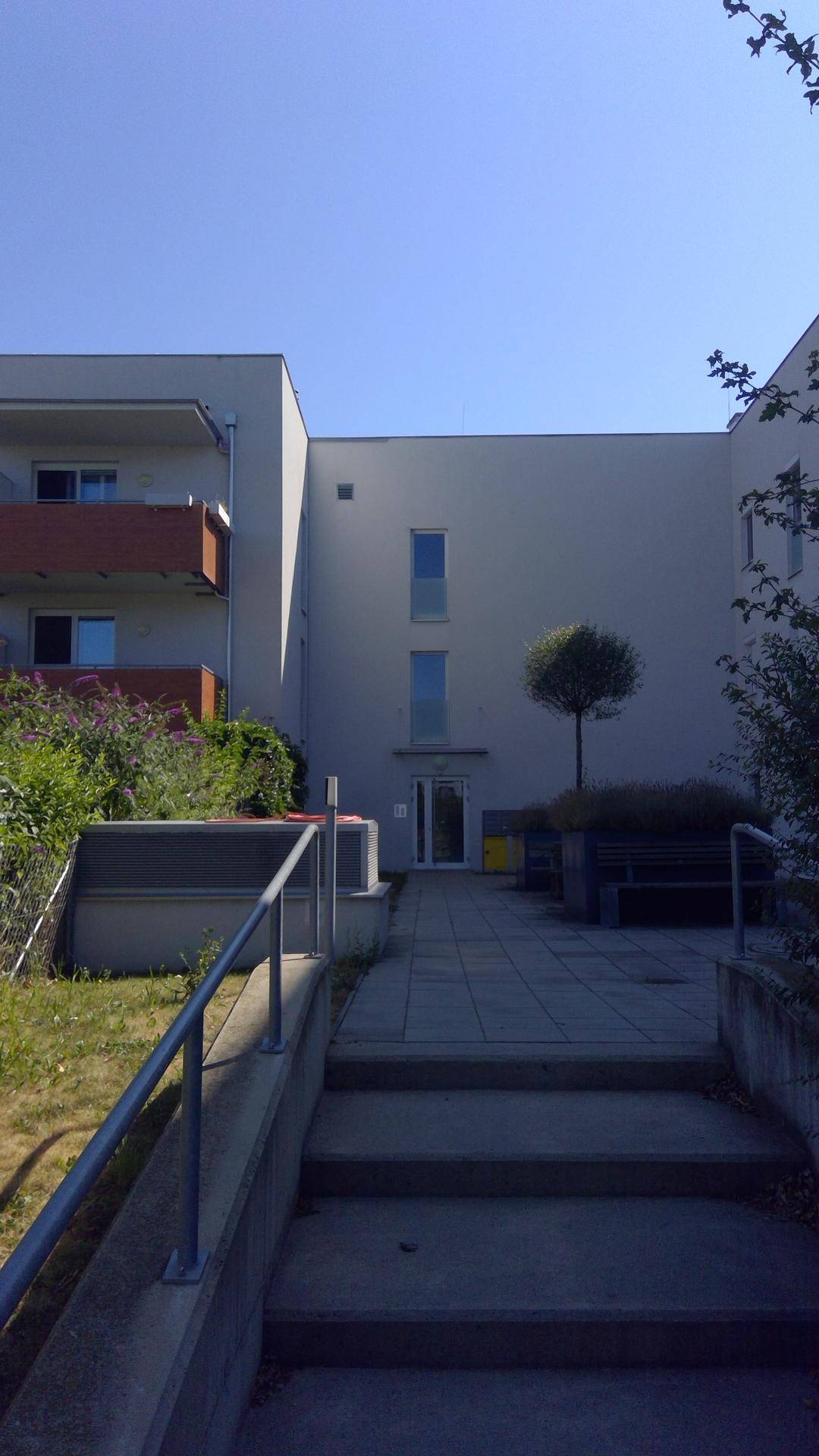 Außenansicht des modernen Gebäudes mit Außentreppe und gepflastertem Vorplatz unter klarem blauem Himmel.
