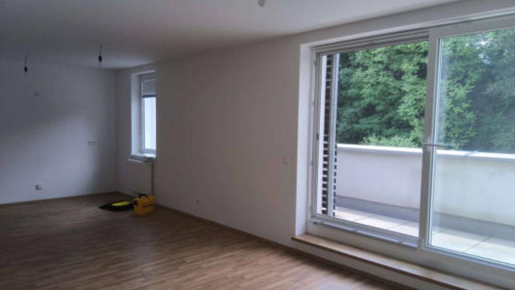 Geräumiger, heller Raum mit Holzboden und Zugang zu einem Balkon, umgeben von viel Grün.