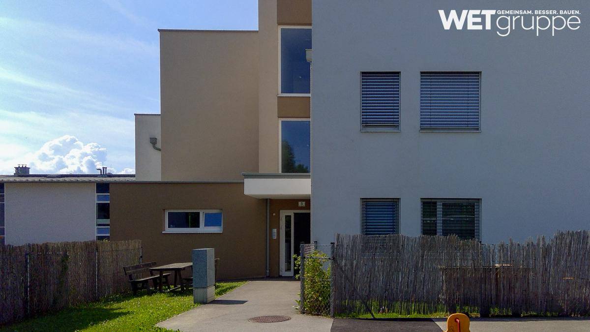 Eingangsbereich eines modernen Wohnhauses mit kleinem Vorgarten, Sitzbank und Jalousien an den Fenstern.