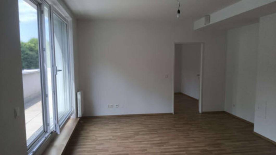 Heller, leerer Raum mit Holzboden und großer Glasfront, die zu einem Außenbereich führt.