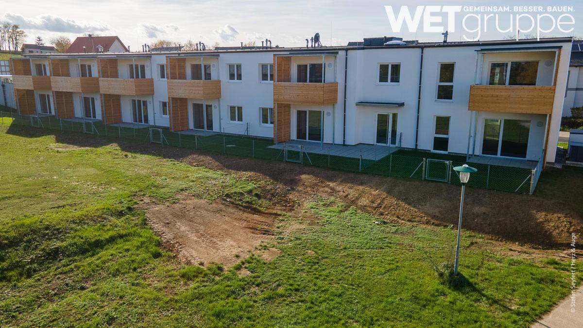 Reihenhäuser mit Holzbalkonen und Gärten in einer grünen Umgebung.