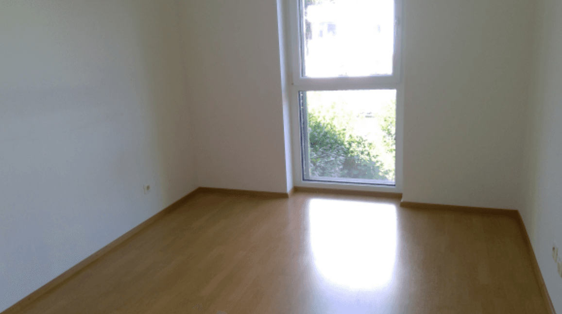 Leeres Zimmer mit Laminatboden und einem großen Fenster, das viel Tageslicht hereinlässt.
