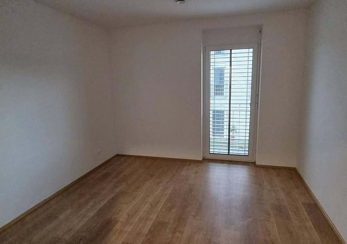 Leeres Zimmer mit Laminatboden und großem Fenster, das viel Tageslicht hereinlässt.