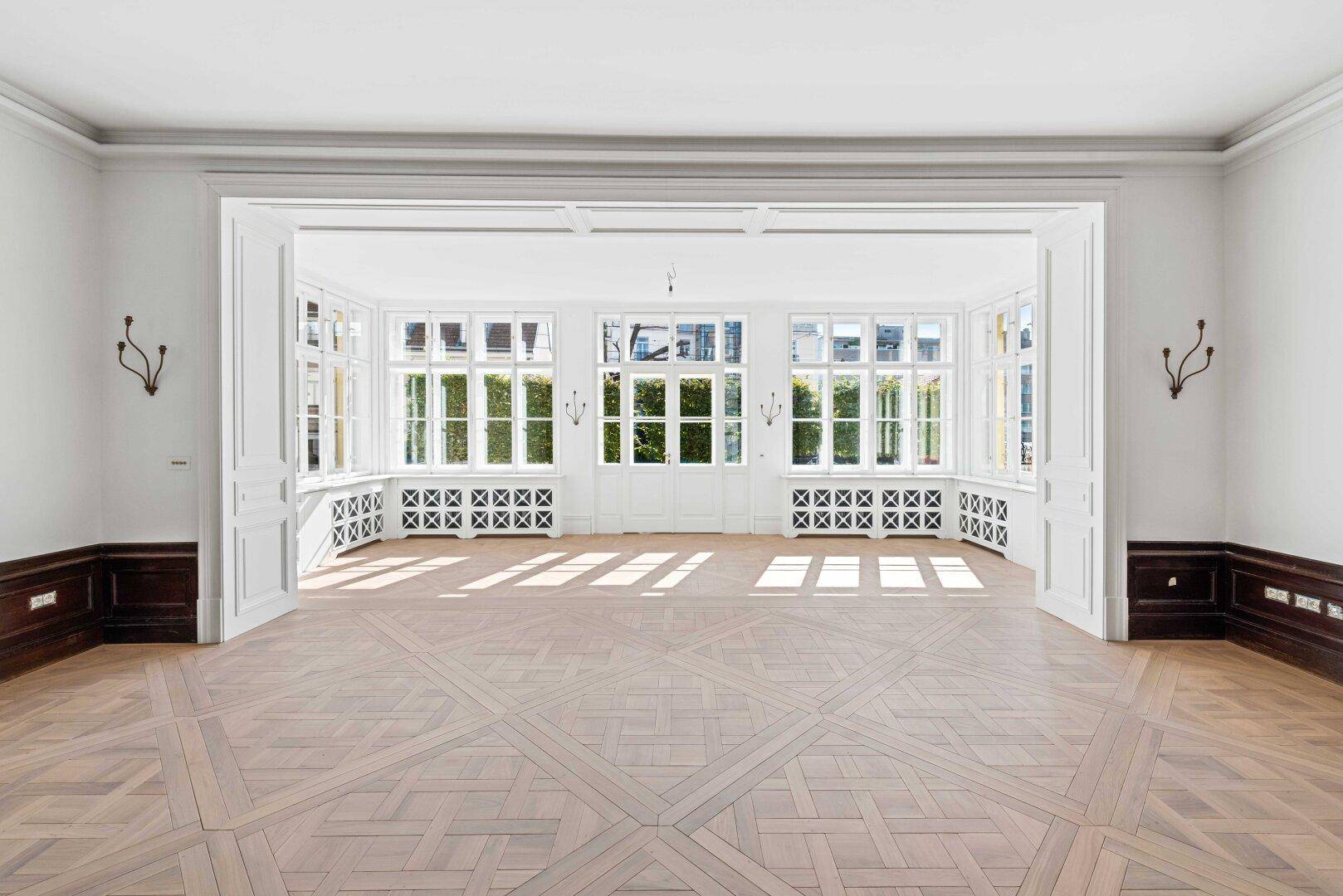 Heller, leerer Raum mit elegantem Parkettboden und großen Fenstern mit Blick ins Grüne.