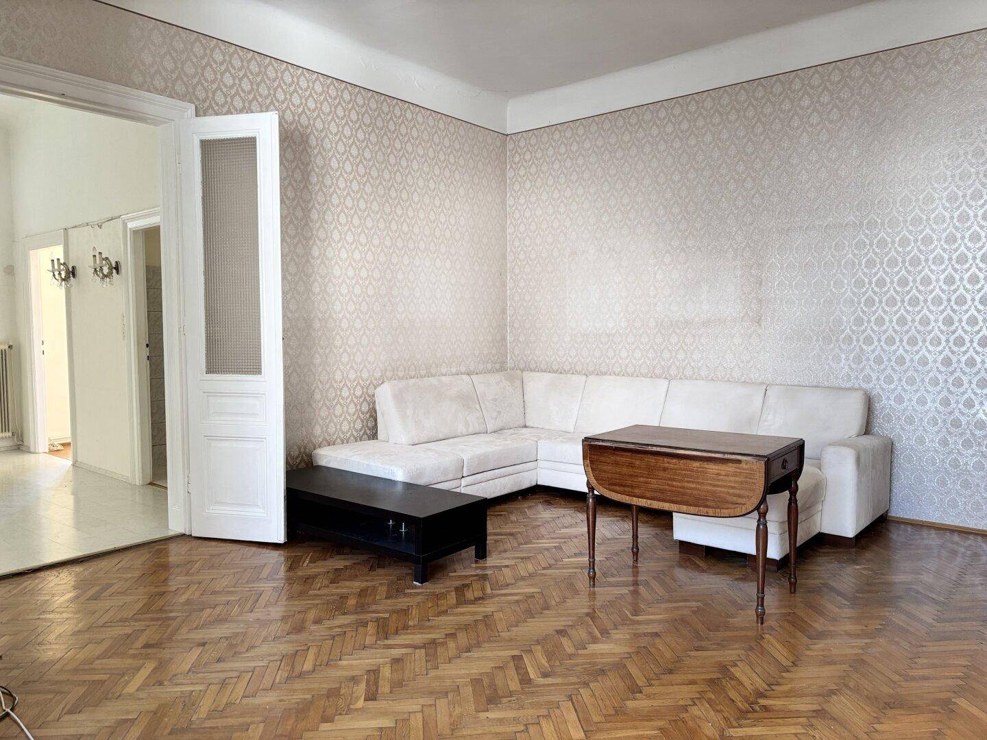 Wohnzimmer mit weißem Ecksofa, Holztisch und Fischgrätparkett, die Wände sind tapeziert.