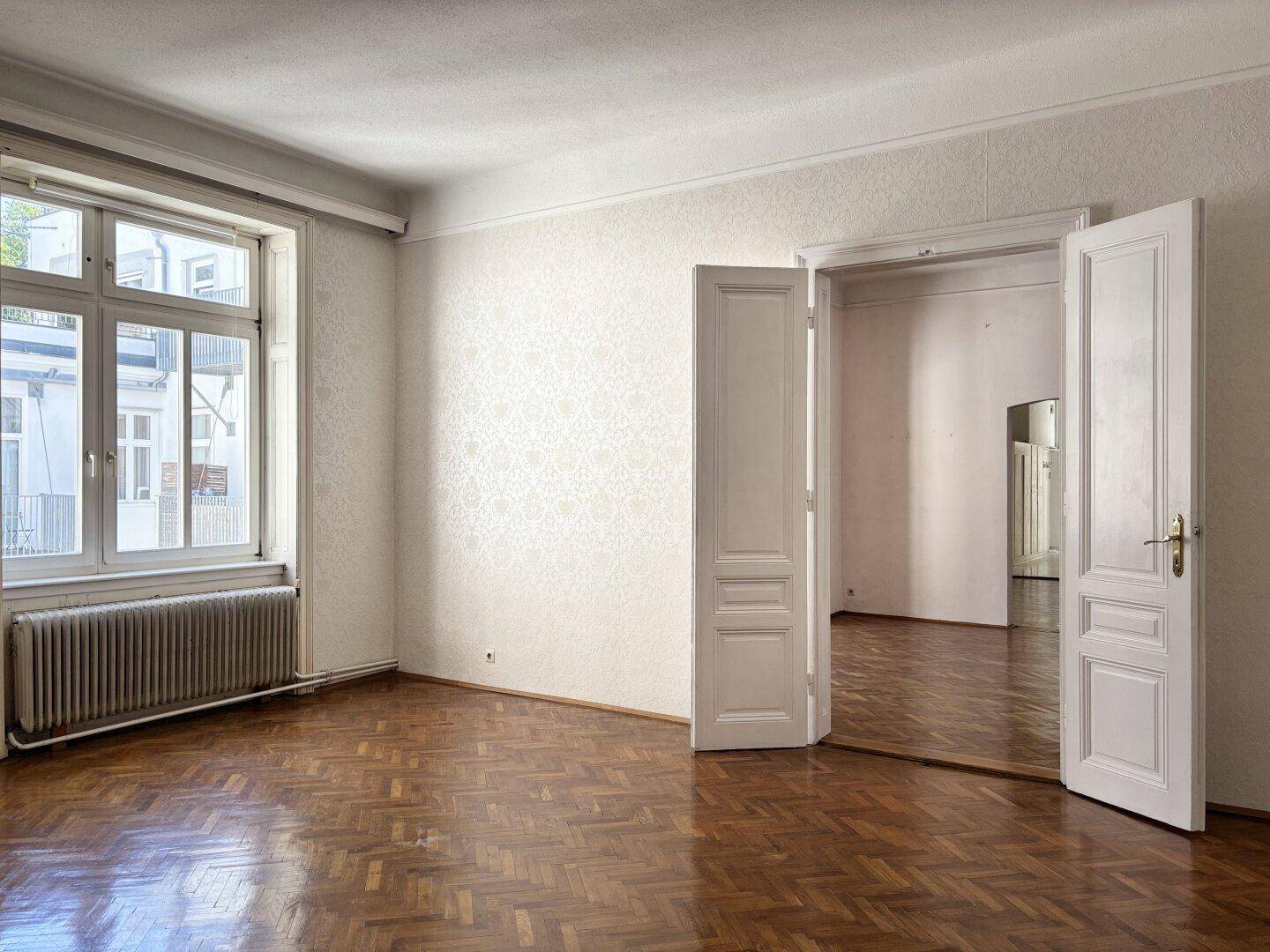Leeres Zimmer mit Fischgrätparkett, großem Fenster, Heizkörper und Doppeltüren zu einem weiteren Raum.