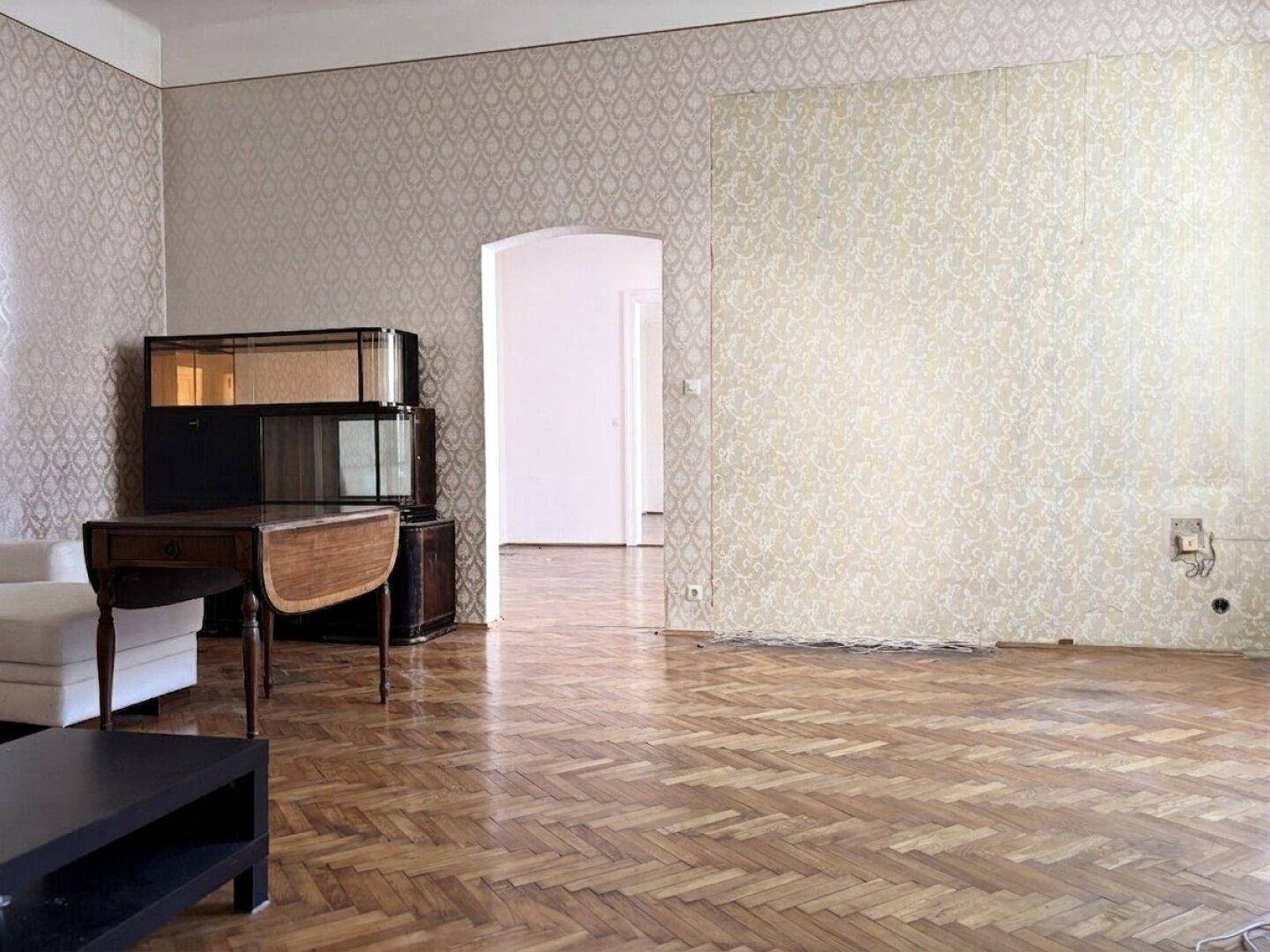 Geräumiges Wohnzimmer in einem Altbau mit Fischgrätparkettboden, gemusterter Tapete und hohen Decken.