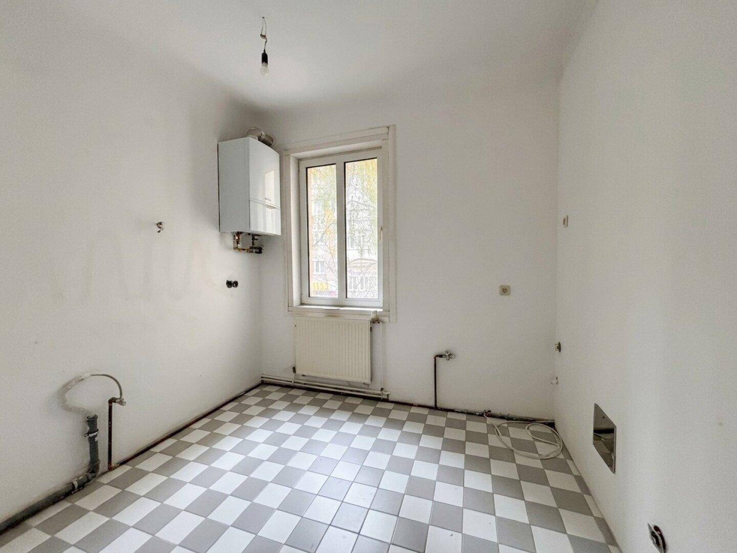 Leere Küche mit karierten Fliesen, Fenster und sichtbaren Anschlüssen für die Kücheninstallation.