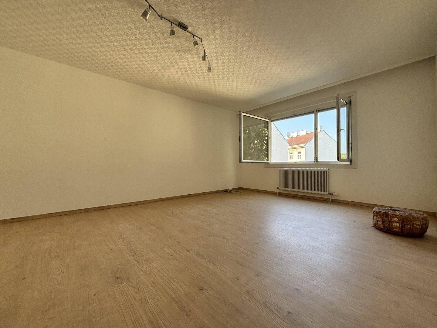 Leeres Wohnzimmer mit Laminatboden, Heizkörper und Blick auf die umliegenden Gebäude.