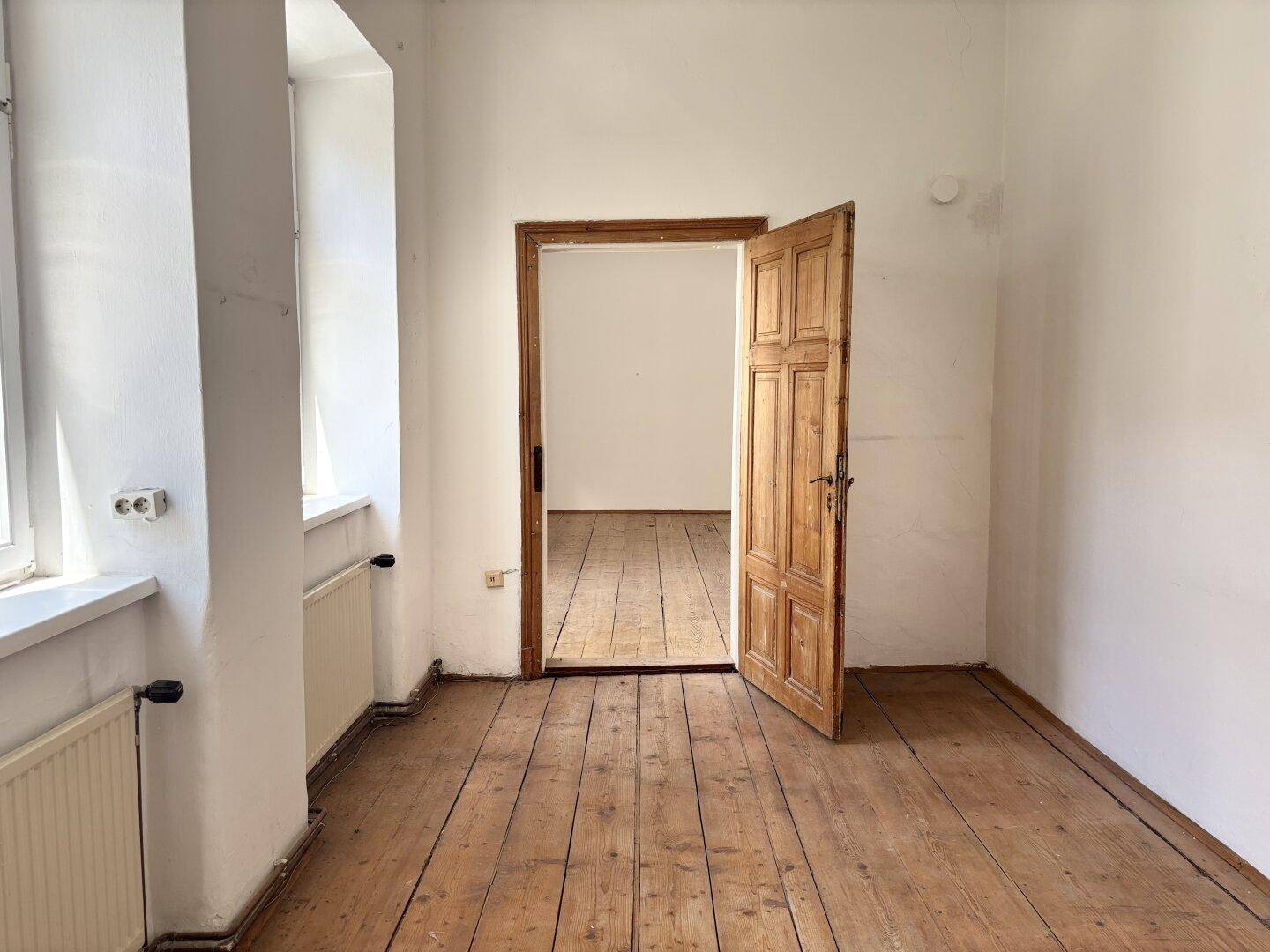 Leeres Zimmer in einem Altbau mit Holzdielenboden, weißen Wänden und Fenstern, die viel Tageslicht hereinlassen.