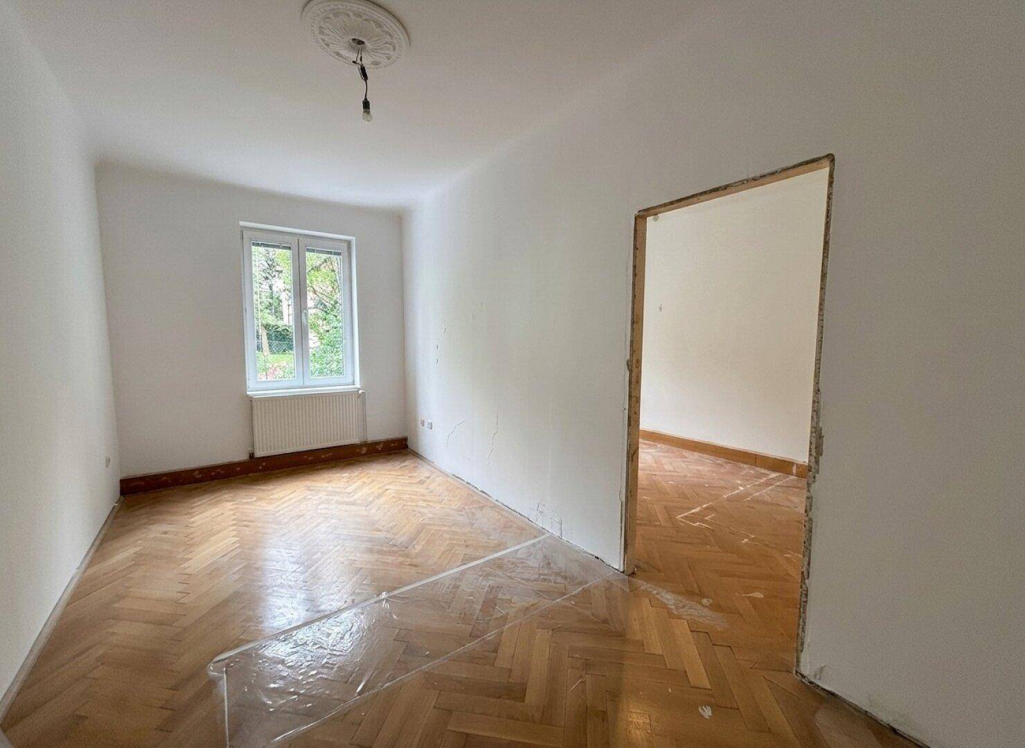 Helles Zimmer mit Fischgrätparkett, Stuckrosette an der Decke und Zugang zu einem weiteren Raum.