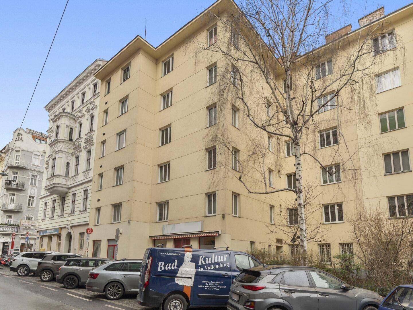 Mehrstöckiges Wohngebäude mit gelber Fassade und parkenden Autos an einer städtischen Straße.