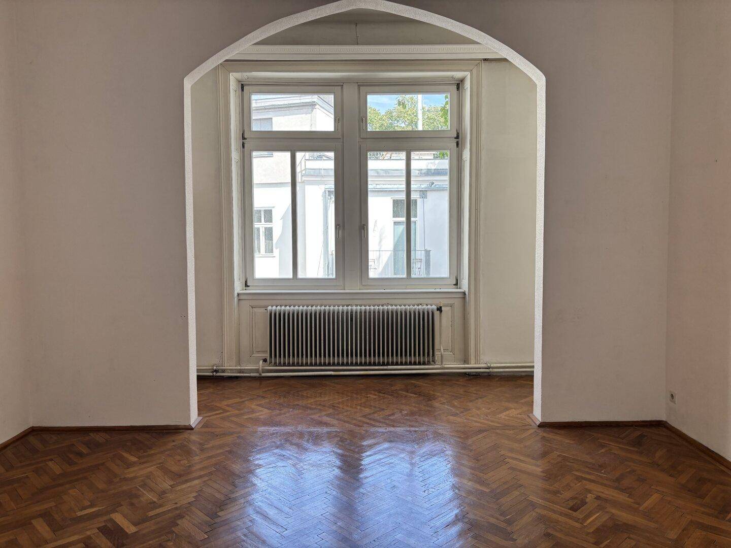 Heller Raum mit Fischgrätparkett, großen Fenstern und einem Heizkörper unter einem eleganten Bogen.