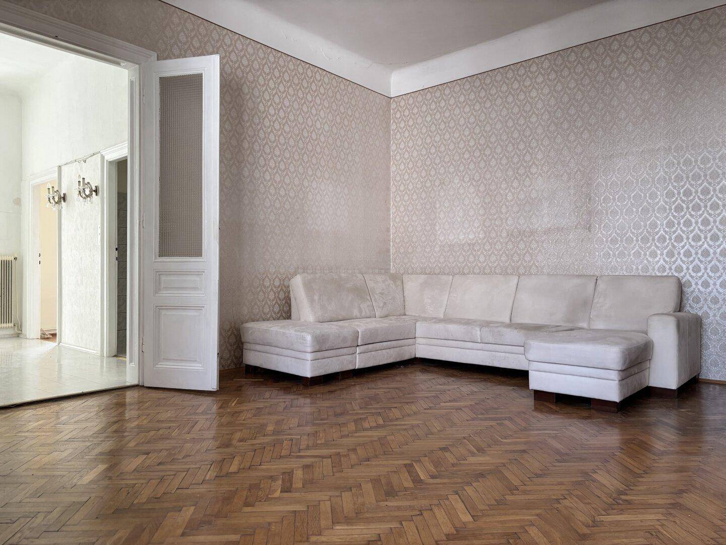Gemütliches Wohnzimmer mit hellem Ecksofa und glänzendem Fischgrätparkett, durch eine Tür verbunden.