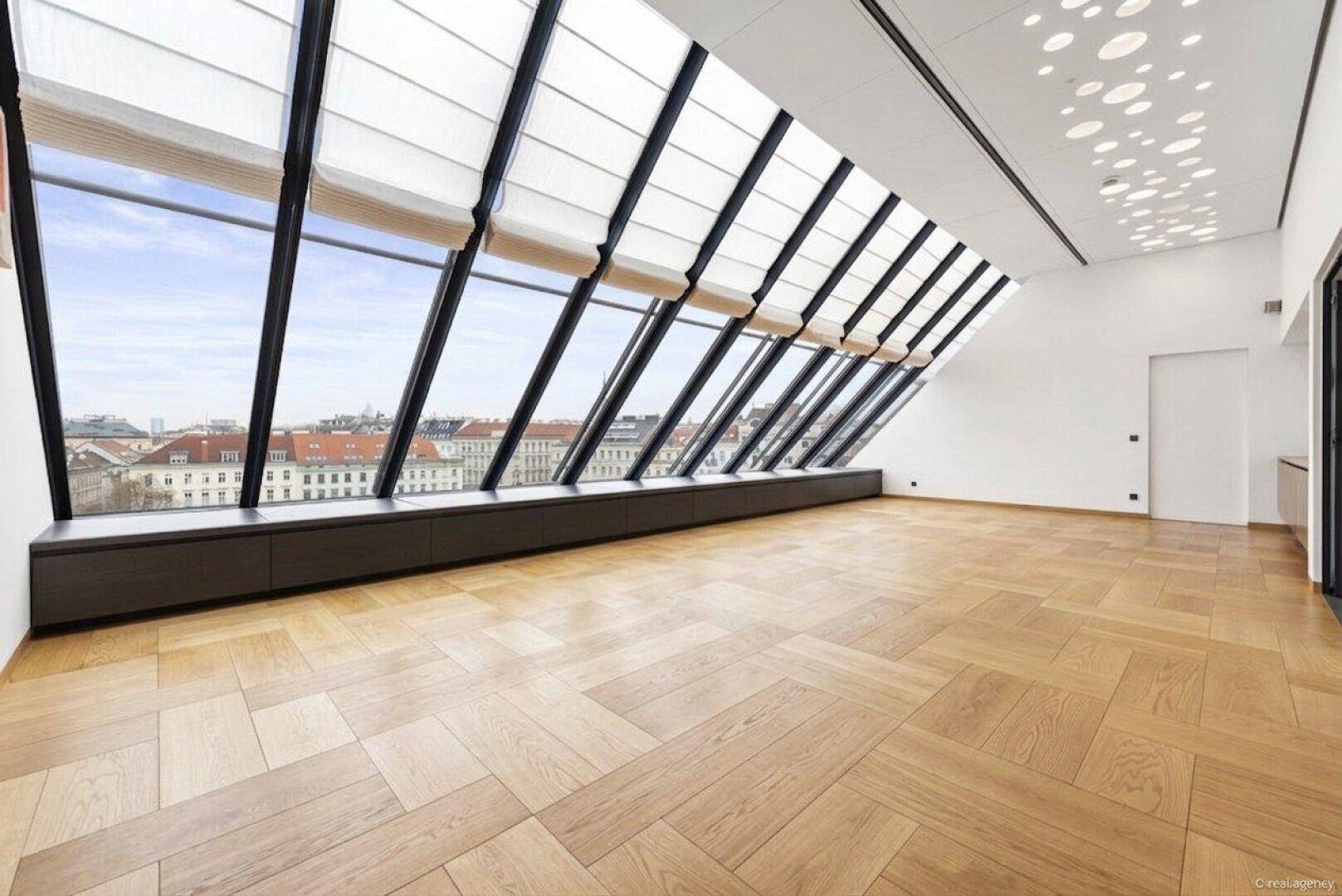 Heller Raum mit schrägen Panoramafenstern, Parkettboden und weitem Stadtblick.