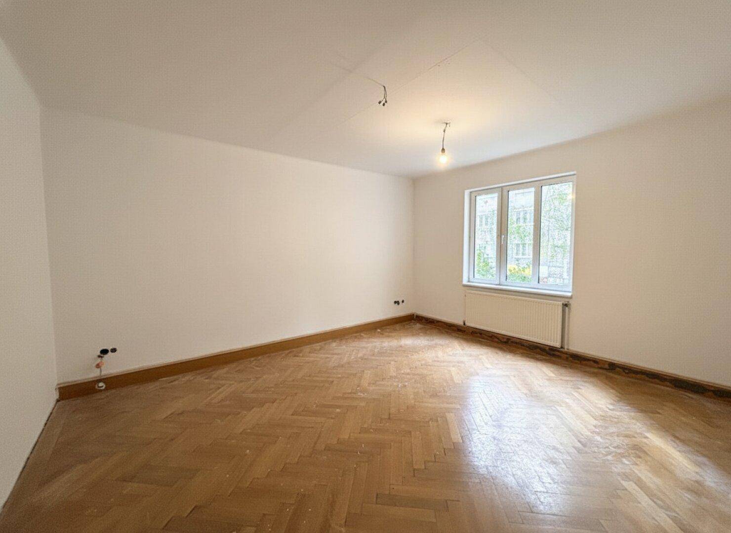 Geräumiges, leeres Zimmer mit hellem Fischgrätparkett und einem Fenster, das viel Tageslicht hereinlässt.