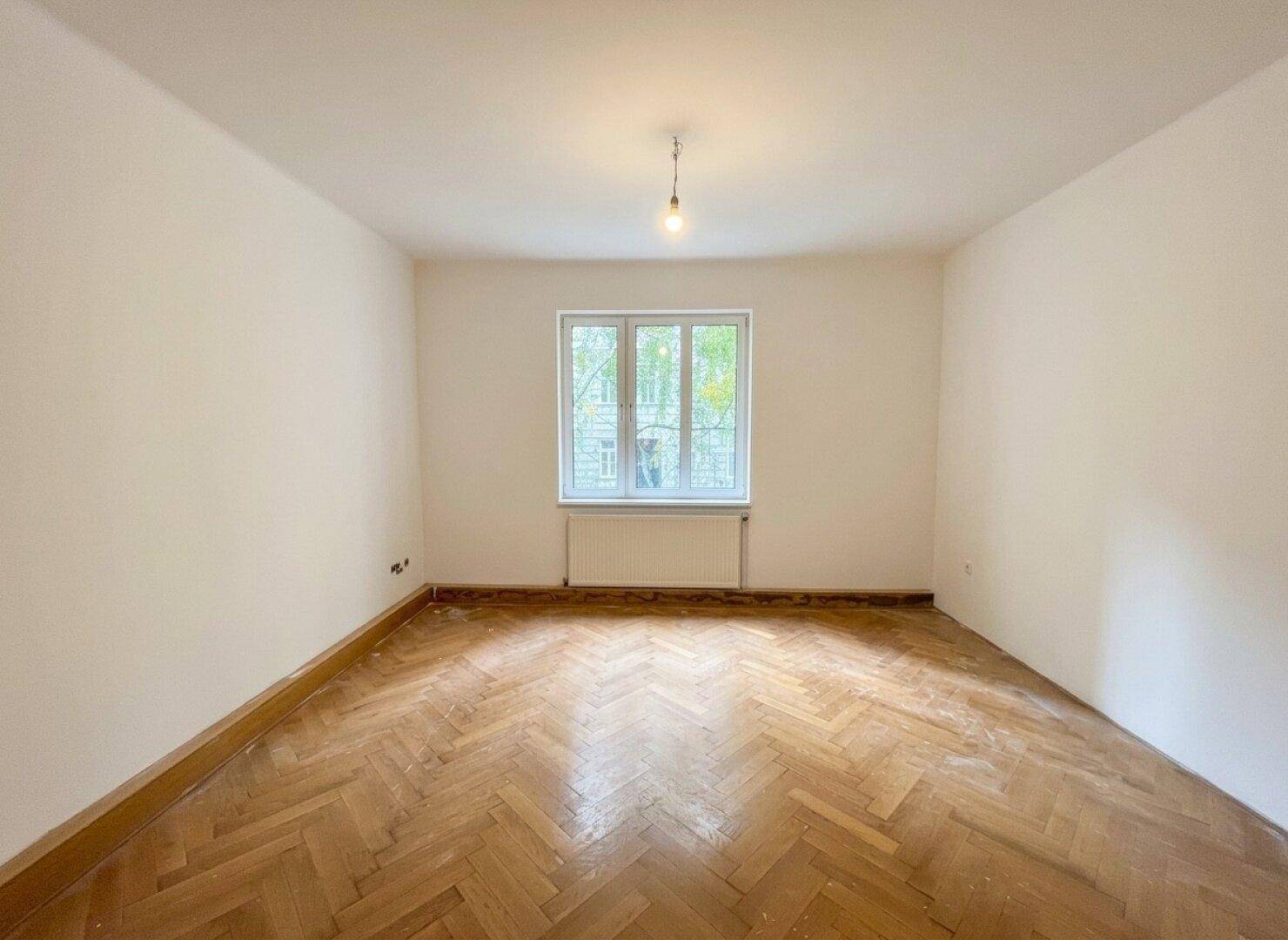 Leeres Zimmer mit klassischem Fischgrätparkett und einem großen Fenster für optimale Belichtung.
