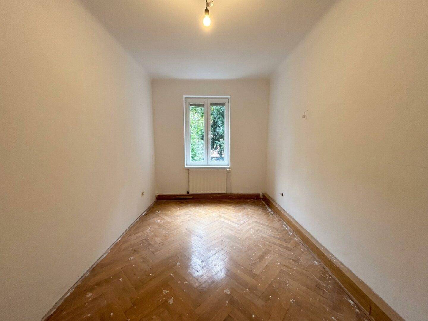 Geräumiger, leerer Raum mit Fischgrätparkett und einem Fenster, das eine angenehme Helligkeit bietet.