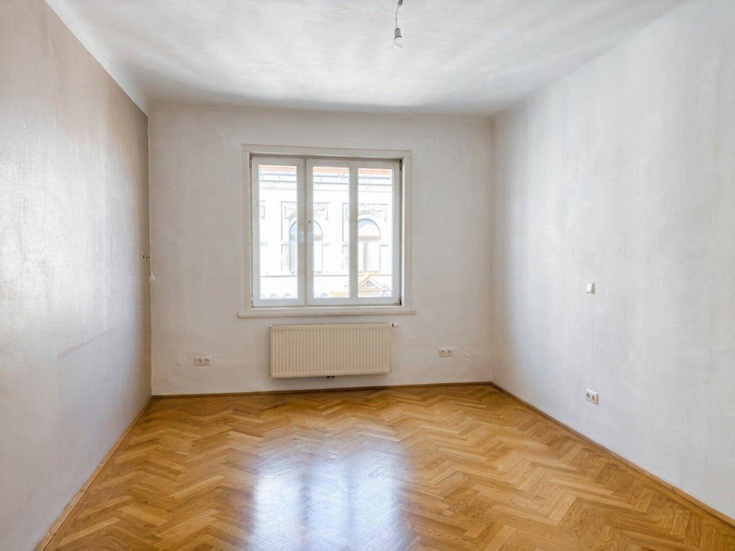 Helles Zimmer mit Fischgrätparkettboden und einem Fenster, das viel Tageslicht hereinlässt.