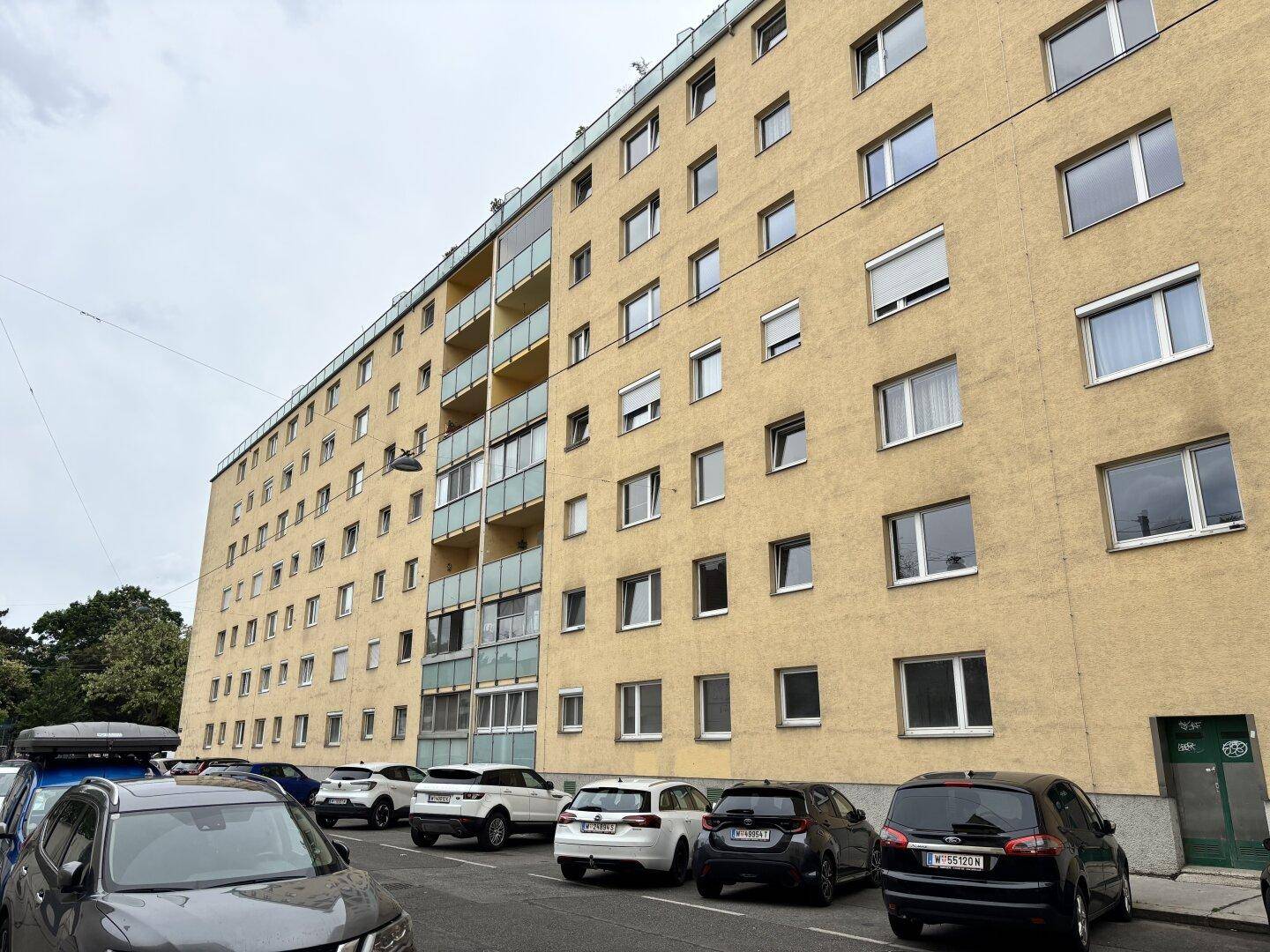 Mehrstöckiges Wohngebäude mit gelber Fassade, zahlreichen Fenstern und Balkonen an einer Straße mit Parkplätzen.