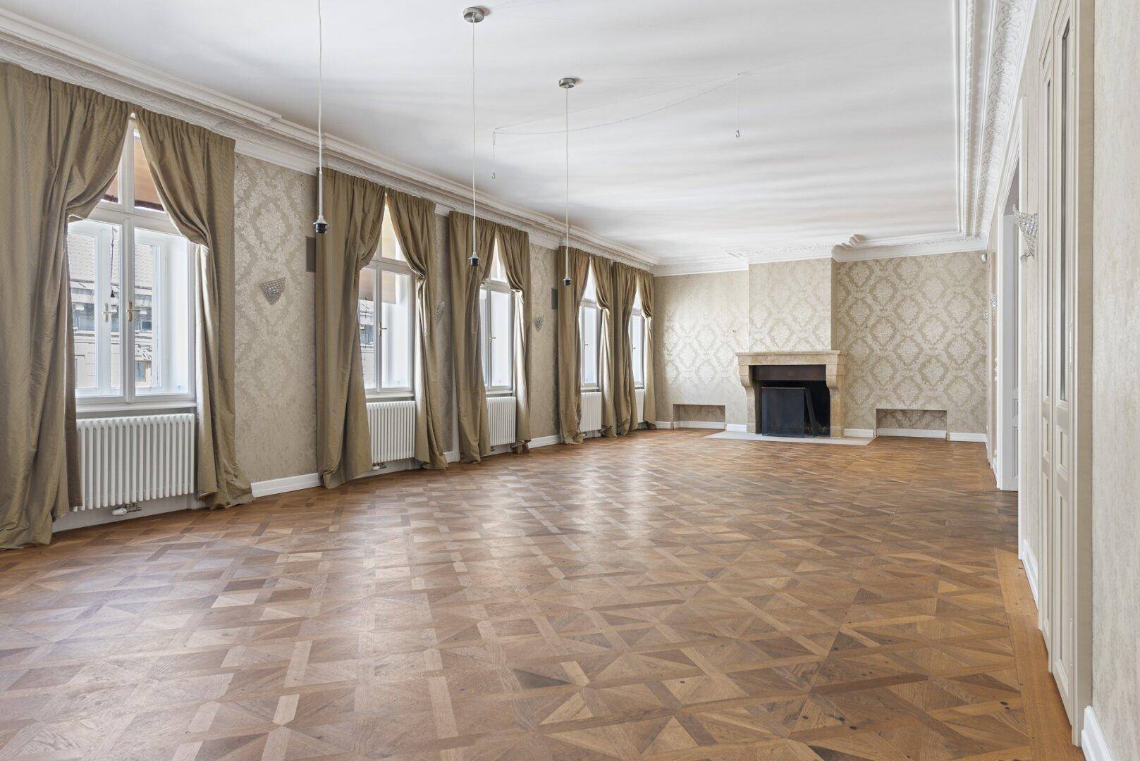 Geräumiges Wohnzimmer mit elegantem Parkettboden, hohen Fenstern und einem klassischen Kamin.