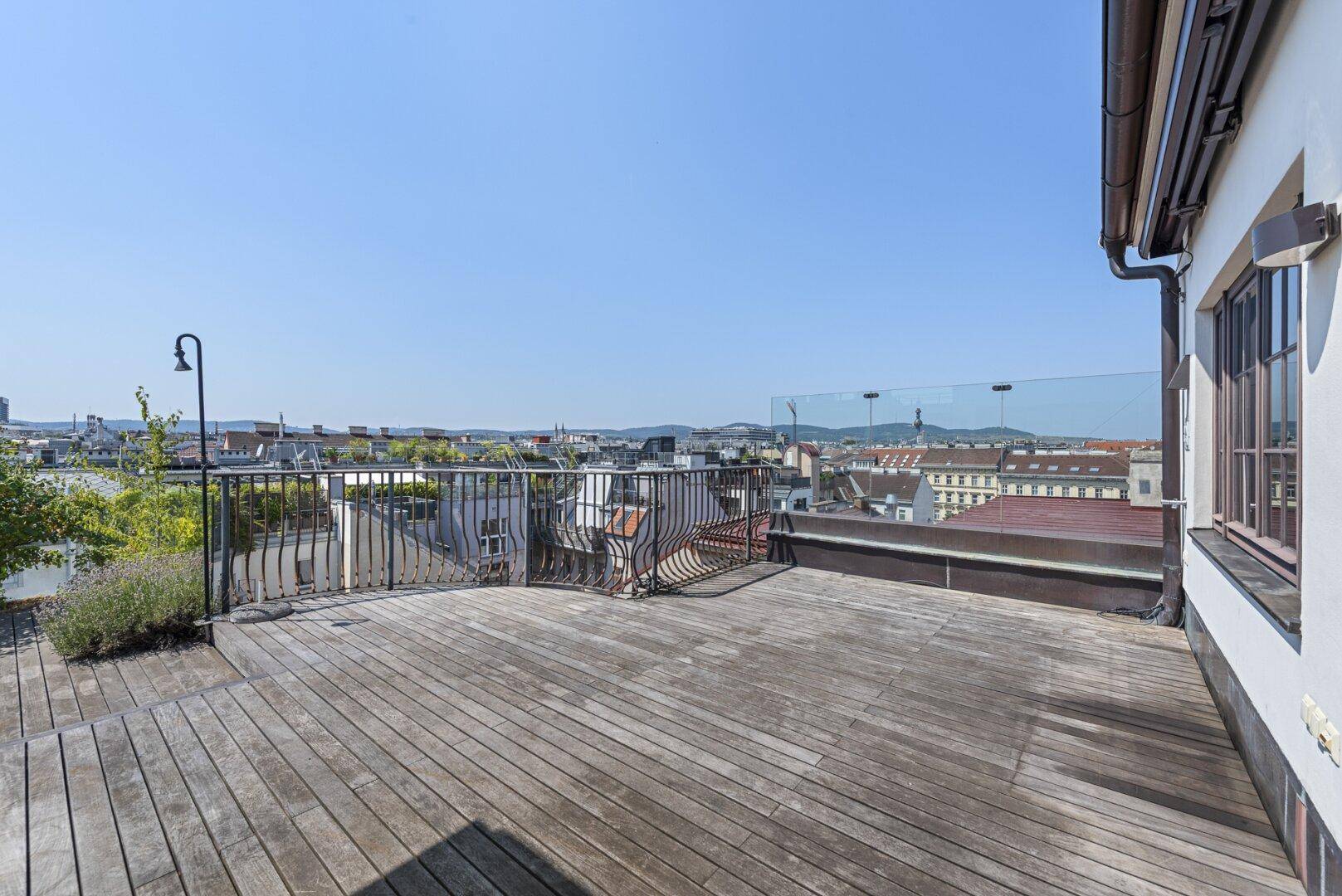 Geräumige Dachterrasse mit Holzbelag, Glasgeländer und weitem Blick über die Stadt.