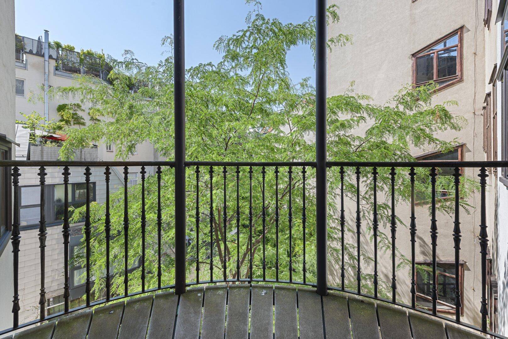 Kleiner, geschwungener Balkon mit Blick auf einen begrünten Innenhof und alte Bäume.