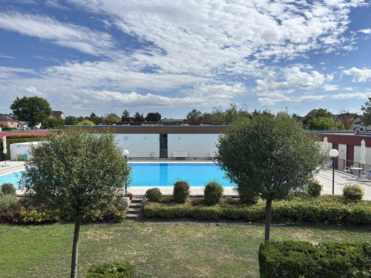 Großer Außenpoolbereich mit klarem Wasser, umgeben von gepflegtem Rasen und Bäumen unter blauem Himmel.