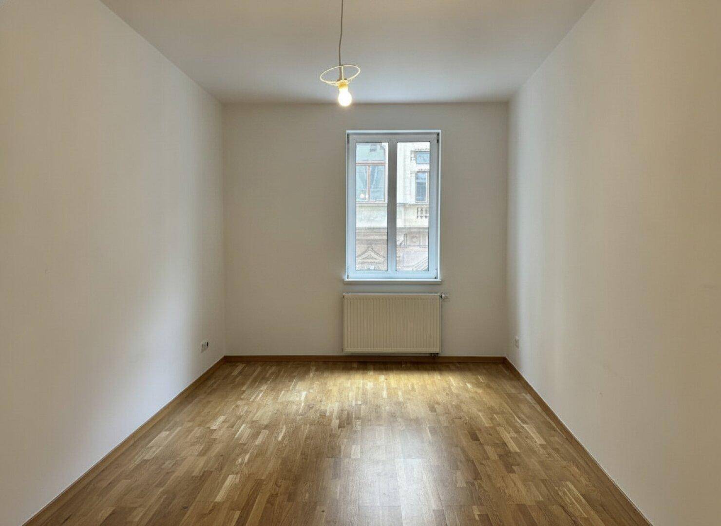 Helles Zimmer mit Holzboden, weiß gestrichenen Wänden und Blick auf die gegenüberliegende Fassade.