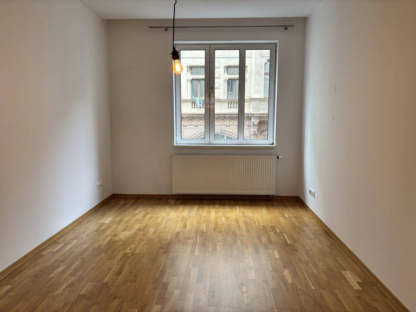 Geräumiges Zimmer mit Parkettboden und großem Fenster, das viel Tageslicht hereinlässt.